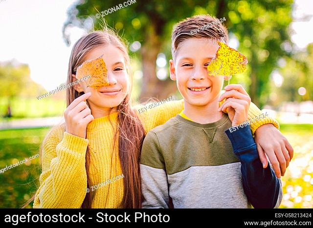 Happy twins teenagers boy and girl posing hugging each other in autumn park holding fallen yellow leaves in hand in sunny weather. Autumn season theme