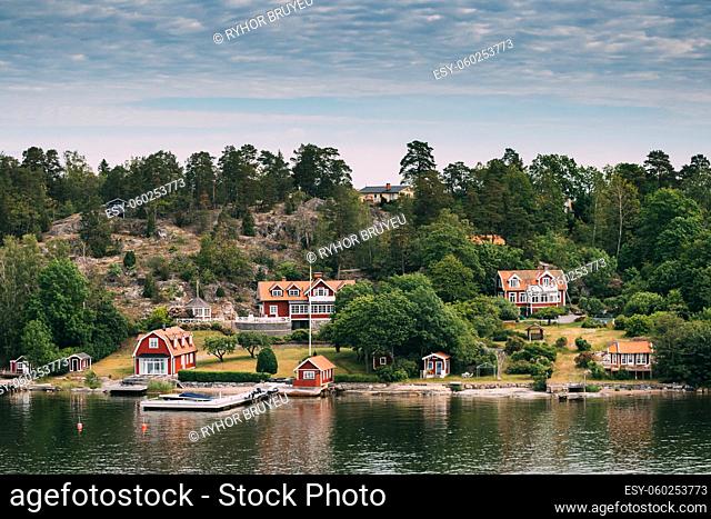 Sweden. Many Beautiful Red Swedish Wooden Log Cabins Houses On Rocky Island Coast In Summer Sunny Evening. Lake Or River Landscape