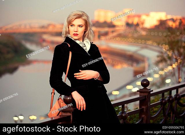 Fashion blond woman in black coat with handbag leaning on fence outdoor