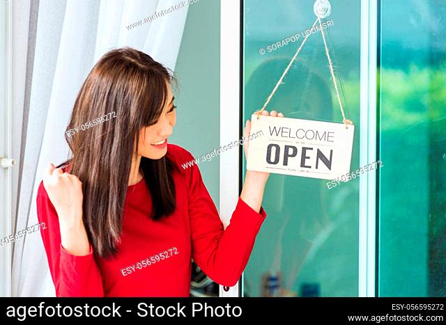 Asian young woman glad and smiling she notice sign wood board label ""WELCOME OPEN"" hanging through glass door front shop