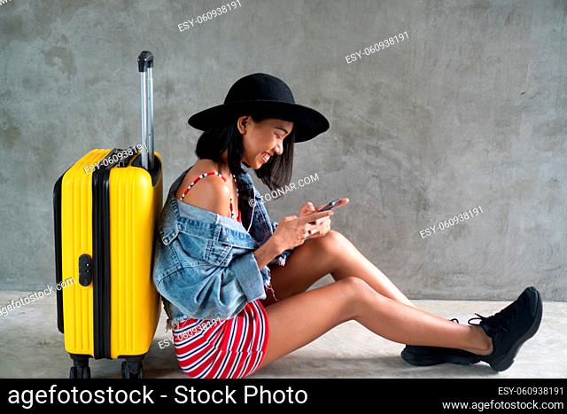 Portrait of smiling young Asian woman with luggage and mobile phone isolated over concrete wall. People lifestyle, travel concept