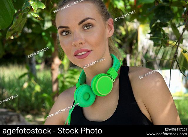Close up of young pretty fitness female wearing bright green headphones smiling and looking into the camera on the beach and enjoying beautiful summer day