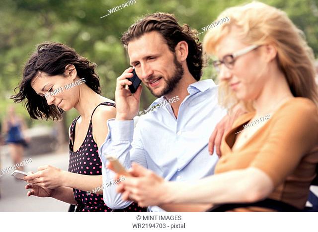 A man and two women seated on a bench in a park, checking their phones, one making a call