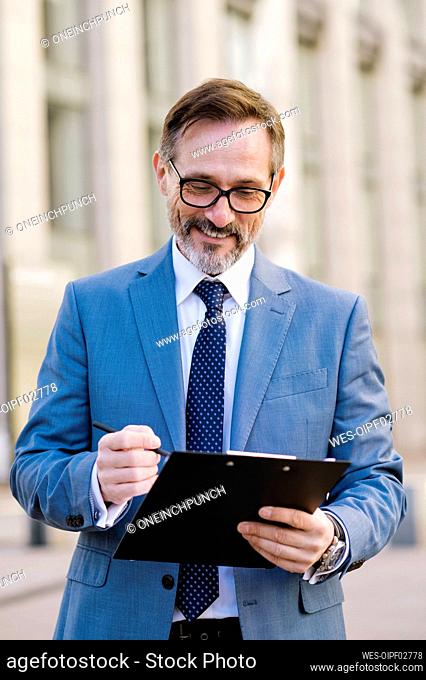 Smiling mature businessman standing with clipboard outside building