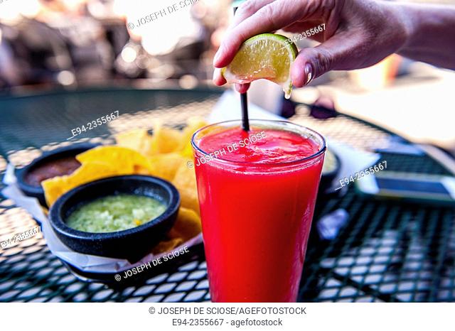 Frozen Strawberry daiquiri on a table at an outdoor restaurant with a woman's hand squeezing a lemon on top of the glass