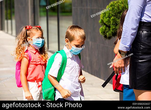 The children stand in protective masks and backpacks in front of the school entrance. The teacher checks the students for compliance with safety rules
