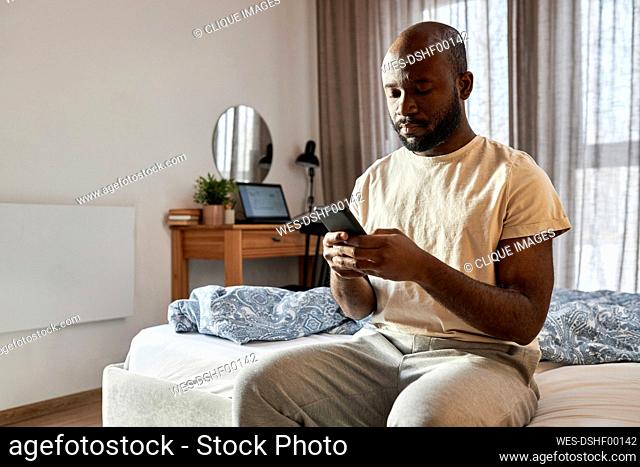 Man using smart phone sitting on bed in bedroom at home