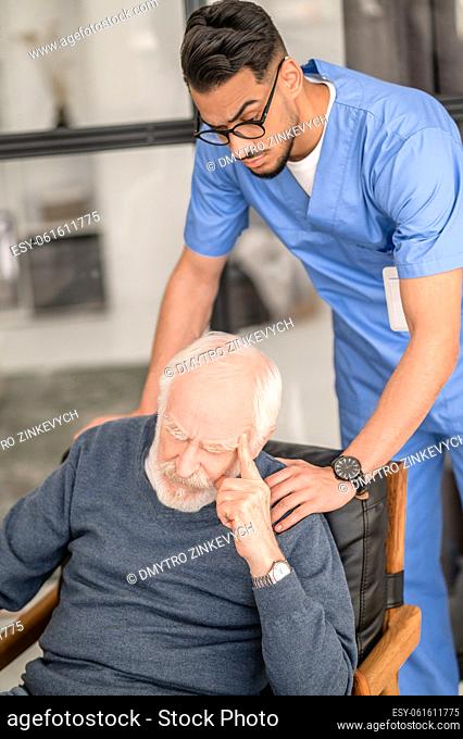 Serious young in-home male nurse in uniform patting a senior male patient on the shoulder