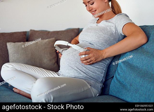 Smiling happy pregnant young woman providing music with earphones to her unborn baby by holding a set of stereo headphones to her abdomen as she sits cross...