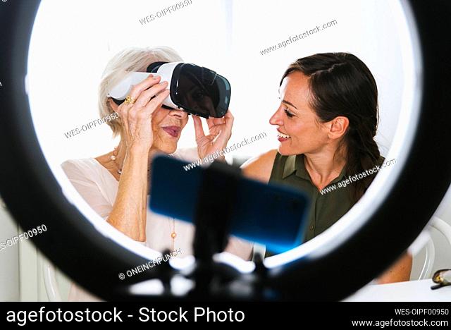 Smiling woman looking at grandmother wearing virtual reality simulator while vlogging at home