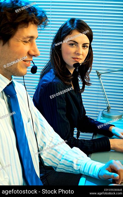 Two young customer service operators sitting at desk, talking on headset, smiling