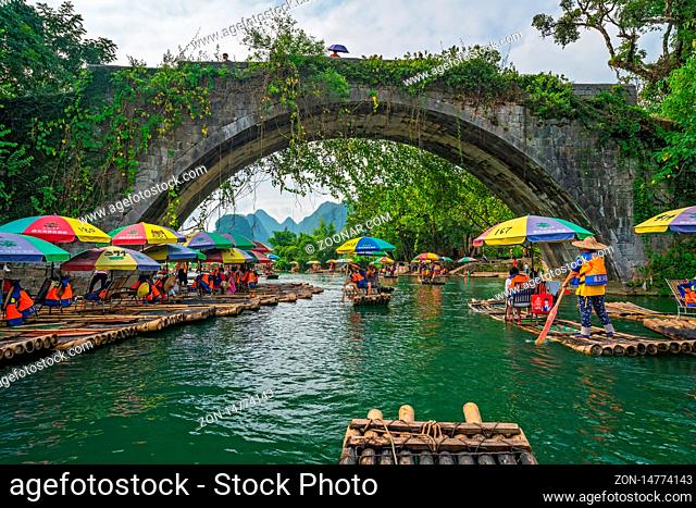 Yangshuo, China - August 2019 : Small local tourist passenger bamboo boats carrying people under the historic Yulong Qiao bridge on the trip along the scenic...