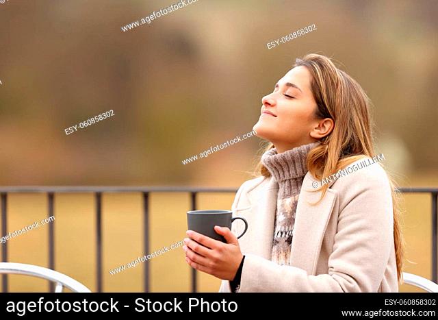 Happy woman holding coffee mug and breathing fresh air in winter in a terrace