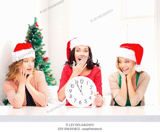 winter, holidays, time and people concept - smiling women in santa helper hats with clock over living room and christmas tree background
