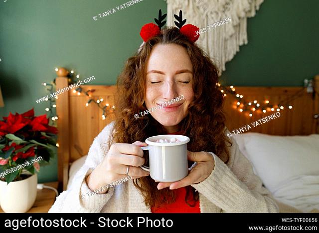 Smiling woman with eyes closed holding cup of hot chocolate on bed at home