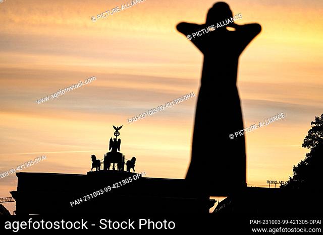 03 October 2023, Berlin: The clouds behind the Brandenburg Gate and the statue ""The Caller"" by Gerhard Marcks are brightly lit on the morning of the...