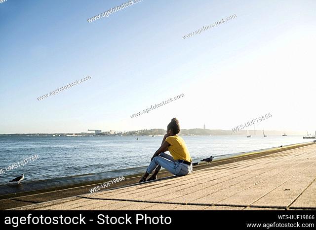 Rear view of young woman sitting at the waterfront at sunset, Lisbon, Portugal
