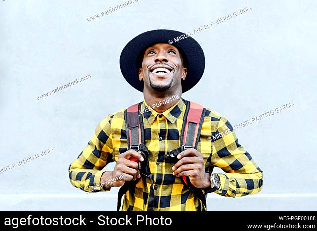 Smiling african man with backpack in hat looking up against white wall