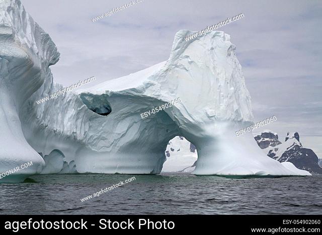iceberg with an arch in Antarctic waters against the backdrop of the mountains of the Antarctic Peninsula on a cloudy day