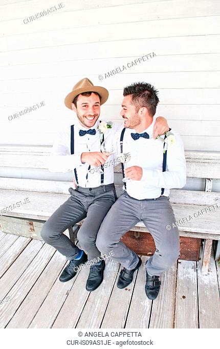 Portrait of groom sitting on bench with male friend, clinking glasses, smiling