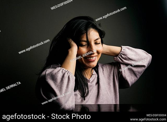 Smiling woman with hands in hair sitting at table against wall