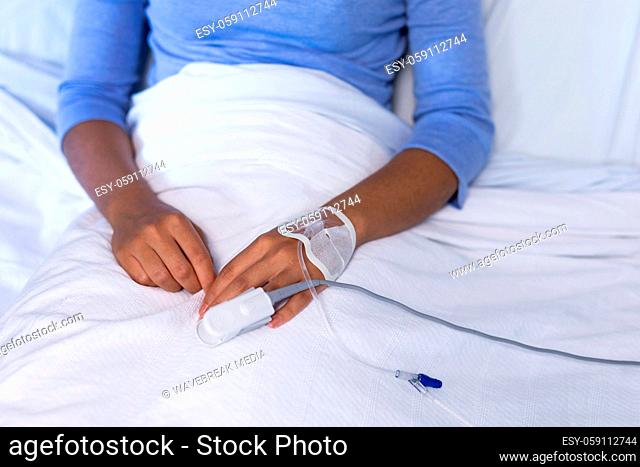 Midsection of mixed race female patient in hospital bed wearing fingertip pulse oximeter and iv tube