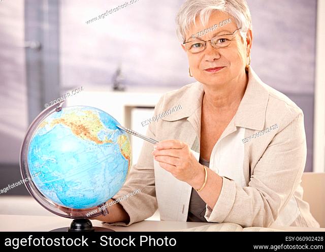 Senior teacher sitting at desk, pointing at globe, teaching geography in elementary school