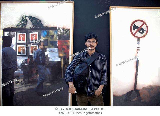 Artist painter Pradeep Mishra standing near his work as reflection of passing pedestrian ; Kala ghoda festival annual art fair ; Bombay Mumbai ; Maharashtra ;...