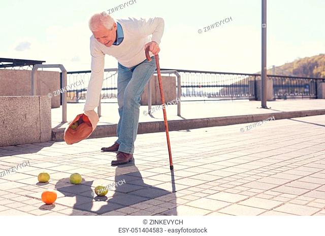Disappointing accident. White-haired elderly man with a cane dropping a bag of fruit and scattering apples and oranges on the ground