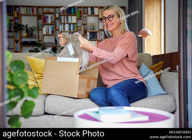 Smiling woman checking clothes in package on sofa at home
