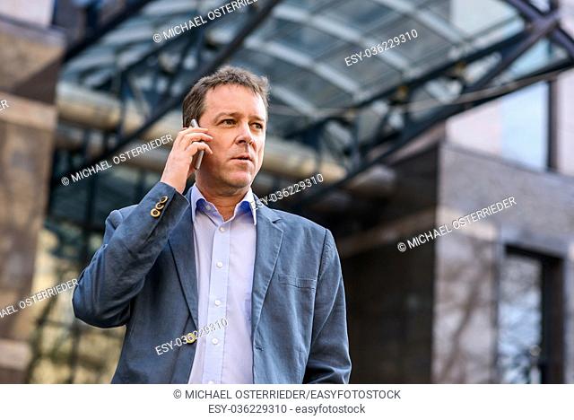 A middle age businessman standing in front of an office building while talking on his phone