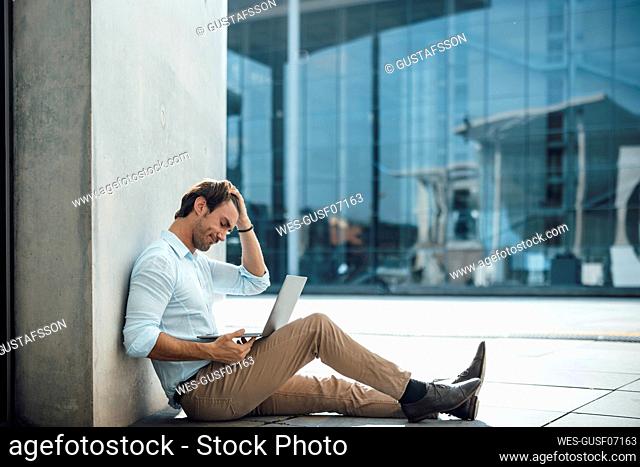 Businessman with hand in hair using laptop by wall outside building