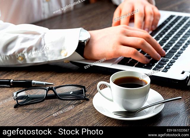Businessman sitting at desk and working with laptop computer. Close-up of man hands typing on keyboard in office. Businessman workplace with glasses and cup of...