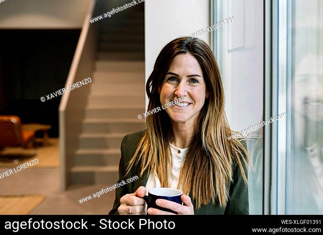 Smiling businesswoman with coffee mug leaning on glass window at office