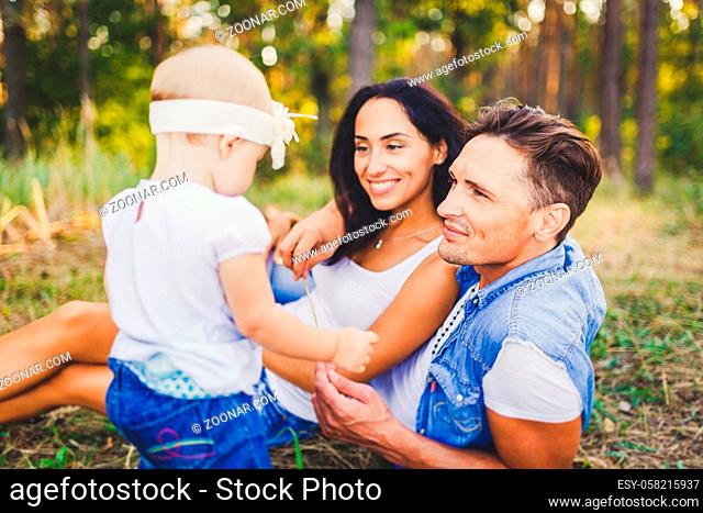 concept of a family vacation in a park in nature. Young stylishly dressed family father and mother lie on the green grass and watch the first steps of the...