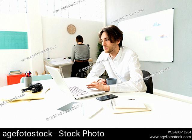 Male entrepreneur using laptop while sitting with colleague in background at office