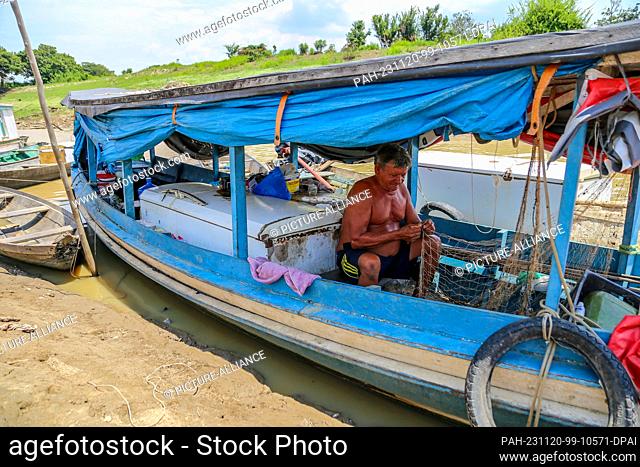 PRODUCTION - 08 November 2023, Brazil, Parintins: A fisherman sits in his boat, almost dry, next to a fridge and mends a net