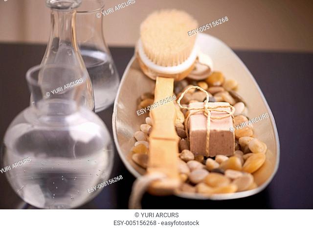 An exfoliating brush, smooth pebbles and a cake of soap beside clear jugs of water