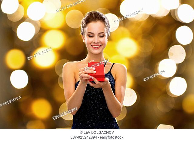 holidays, presents, luxury and people concept - smiling woman in dress holding red gift box over lights background