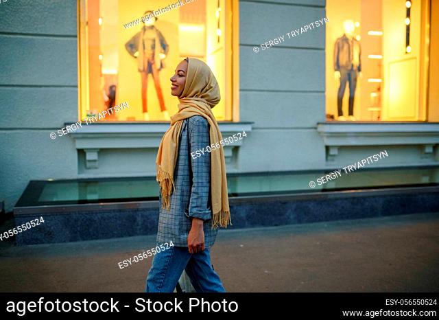 Arab girl in hijab looking on showcase of fashion store in downtown. Muslim woman walking on street. Female student in big city