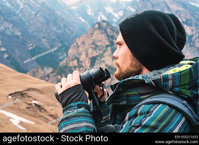 A hipster man with a beard in a hat, a jacket, and a backpack in the caucasian mountains holds binoculars, adventure, tourism, tracking