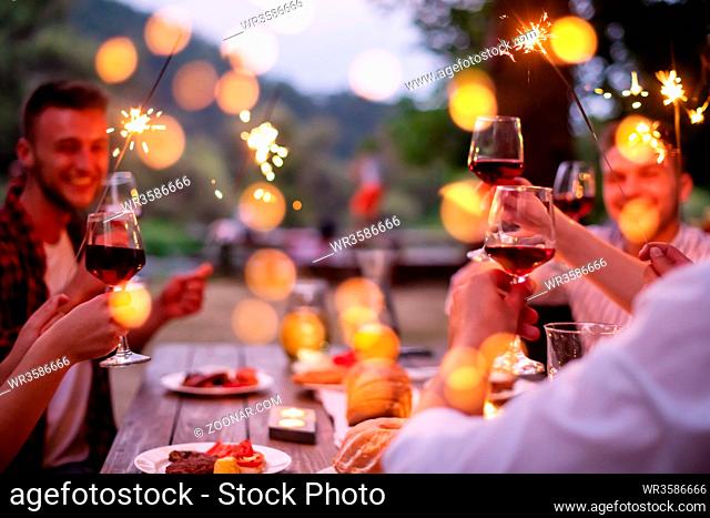 group of happy friends celebrating holiday vacation using sprinklers and drinking red wine while having picnic french dinner party outdoor near the river on...