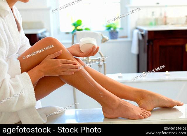 Happy caucasian woman wearing robe, applying cream on legs in bathroom