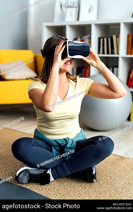 Smiling woman wearing virtual reality simulator sitting with cross-legged in living room