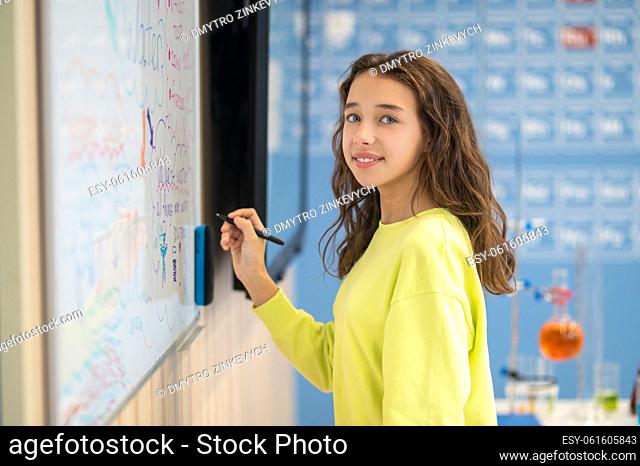 Good mood. Smiling girl with marker in hand standing near blackboard looking at camera in classroom