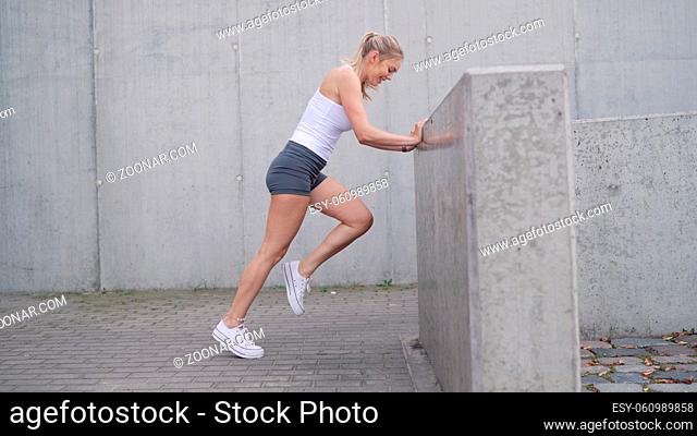 Side view of slim sportswoman in shorts and top leaning on concrete barrier and exercising during fitness training in city