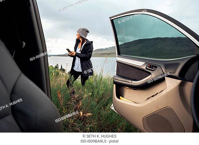 Young woman standing beside Dillon Reservoir, holding smartphone, view through parked car, Silverthorne, Colorado, USA