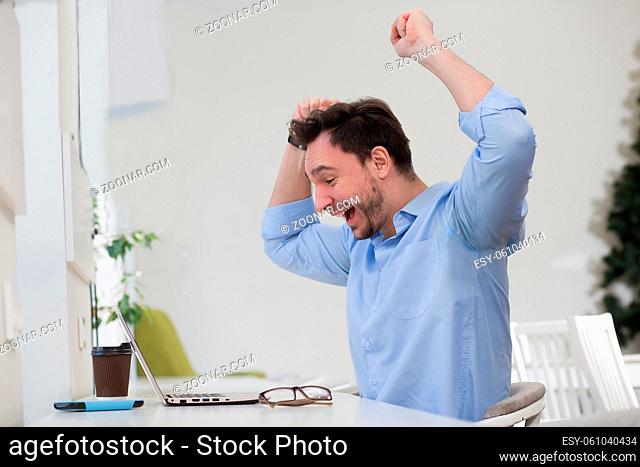 Happy man expressing his positivity with his arms raised. Handsome man in blue shirt looking at laptop computer's screen