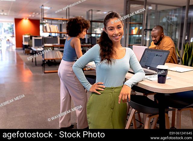 Portrait of smiling young hispanic businesswoman standing by desk against biracial colleagues