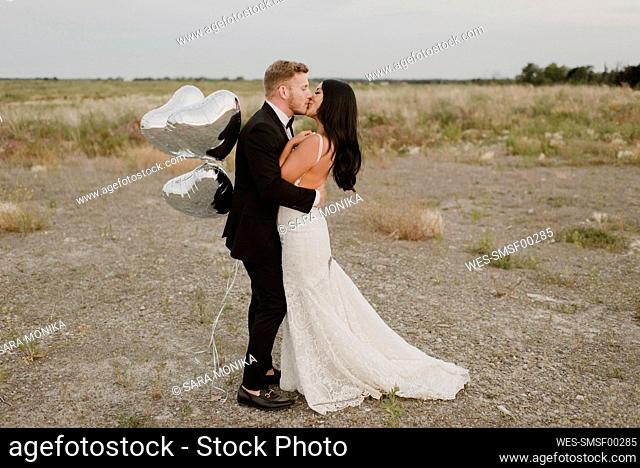 Smiling groom with heart shape balloons kissing bride against sky